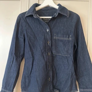 Jeansskjorta från Massimo Dutti - Mörkblå jeansskjorta från Massimo Dutti med klassisk krage, bröstficka och kontrastsömmar. Skjortan har långa ärmar med dubbla knappar vid manschetten och stängs med knappar framtill. Perfekt för en avslappnad men snygg look. Använd 1 gång💕