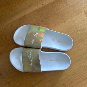 Guldiga sandaler från Tommy Hilfiger - Snygga sandaler från Tommy Hilfiger med vit sula och glansig guldig rem över foten. Remmen har holografisk effekt och logga i vitt. Perfekta för sommardagar och strandhäng.
