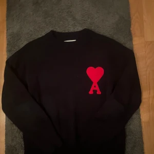 Svart stickad tröja från Ami Paris - Svart stickad tröja från Ami Paris med röd hjärtlogga och bokstaven A på bröstet. Klassisk rund halsringning och långa ärmar. Perfekt för dig som gillar stilrena plagg med en twist. Materialet är mjukt och håller värmen bra.
