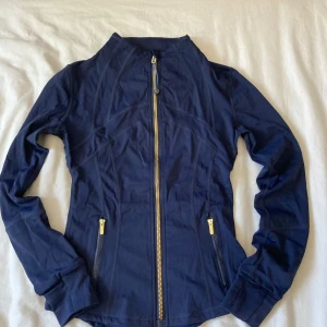 Mörkblå Define Jacket från Lululemon - Säljer en mörkblå Define Jacket från Lululemon med snygg figurnära passform. Jackan har hel dragkedja i guld, två fickor med dragkedja och långärmade ärmar. Materialet är stretchigt och andas, perfekt för träning eller chill. Ikonisk Lululemon-logga på ryggen. Storlek 2 vilket motsvarar S\SX. Helt ny!