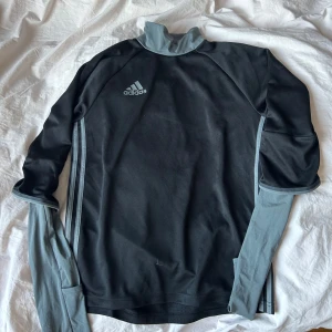 Svart Adidas långärmad träningströja - Svart långärmad träningströja från Adidas med ljusgrå detaljer och klassiska ränder längs sidorna. Hög krage och sportig design, perfekt för träning eller chill dagar. Adidas-logga på bröstet och mjukt material som andas. Aldrig använd.