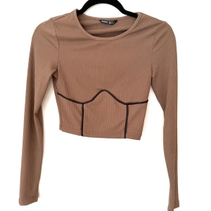 Brun croppad långärmad topp - Säljer en brun croppad långärmad topp i ribbad syntet med svarta kontrastsömmar framtill som ger en cool detalj. Toppen har rund hals och figurnära passform, perfekt till höga jeans eller kjol.