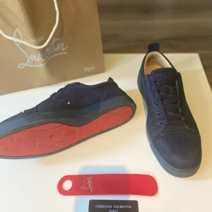Mörkblå sneakers från Christian Louboutin - Snygga mörkblå sneakers från Christian Louboutin i mocka med klassisk röd sula. Skorna har rund tå, platt sula och snörning framtill. Perfekt för dig som vill ha en lyxig och stilren look med en ikonisk touch.