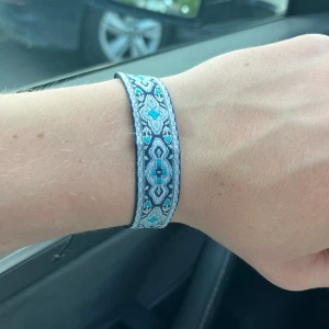 Blått mönstrat tygarmband med snörning - Snyggt armband i tyg med blått och vitt mönster och svarta detaljer. Armbandet har justerbar snörning baktill för perfekt passform. Perfekt accessoar för att ge din outfit lite extra färg och stil.