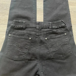 Svarta jeans Dora Comfort - Svarta jeans från Dora i comfort fit med snygga detaljer på bakfickorna i form av broderi och små nitar. Byxorna har resår i midjan och är tillverkade i en mix av bomull, polyester och elastan för extra stretch och bekvämlighet.