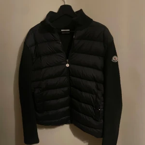Svart moncler cardigan - Svart pufferjacka från Moncler med quiltad framsida och stickade ärmar. Dragkedja framtill, Moncler-logga på ärmen och röd Moncler-text längs sidan. Jackan har en clean och sportig vibe, perfekt för dig som gillar streetstyle. Priset är inte hugget i sten