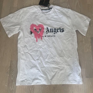 Palm Angels Palm Beach Heart Sprayed Logo T-Shirt - Säljer en vit t-shirt från Palm Angels med svart text och ett stort rosa hjärta med drip-effekt på bröstet. Klassisk passform och rund halsringning. Perfekt statement-plagg för dig som gillar streetwear och unika prints.