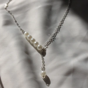 Halsband med pärlor och silverkedja - Stilrent halsband med tunn silverfärgad kedja och fem vita pärlor i rad, samt en extra pärla som hänger i en kort kedja. Perfekt för dig som gillar minimalistiska smycken med en elegant touch. Finns även med guldfärgad kedja.