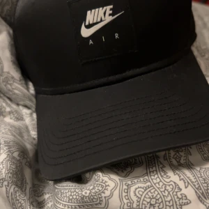 Svart keps från Nike Air - Svart keps från Nike Air med klassisk böjd skärm och justerbar snapback baktill. Kepsen har en tydlig Nike Air-logga framtill i vitt och är tillverkad i ett slätt syntetmaterial. Perfekt för en sportig och stilren look. Kepsen är använd ca 10 gånger och kostar runt 200 ny