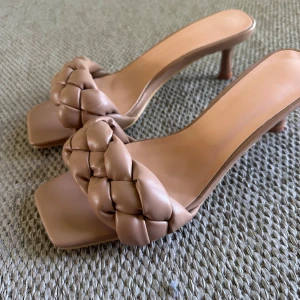 Beige flätade sandaler med klack - Snygga sandaler i beige med flätad rem över foten och fyrkantig tå. Skorna har en smal, låg klack och är tillverkade i ett mjukt, skinnliknande material. Perfekta för dig som gillar trendiga detaljer och stilren design.