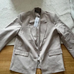 Beige oversize kavaj från Gina Tricot  - Snygg beige kavaj från Gina Tricot med klassisk krage och två knappar framtill. Kavajen har raka fickor med lock och är tillverkad i ett mjukt material som ger en stilren look. Nypris 800kr, helt oanvänd med prislapp kvar🥳