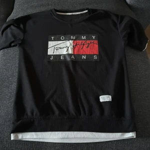 Svart Tommy Jeans t-shirt med logga - Svart t-shirt från Tommy Jeans med stor logga framtill i vitt, rött och svart. Klassisk rund halsringning och korta ärmar. T-shirten har en vit detalj längst ner och är tillverkad i mjukt material som känns skönt mot huden.