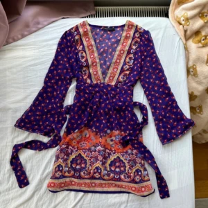 Blommig omlotttopp i lila och orange - Supersöt lång omlotttopp  i lila med blommigt mönster och färgglada detaljer i orange, rött och gult. Djup v-ringning, långa ärmar och knytband i midjan. Perfekt för dig som gillar boho-vibes och färg. Tunn och luftig, passar perfekt till varmare dagar. Toppen är relativt lång och är i ett ganska nytt skick med 0 skavanker. Toppen kan knytas där bak med två band som sitter på varsin sida av toppen