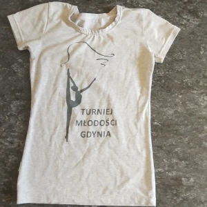 Ljusgrå t-shirt Turniej Młodości - Ljusgrå t-shirt med tryck av en gymnast och texten 'Turniej Młodości Gdynia' på framsidan. T-shirten har rund halsringning och korta ärmar. Materialet känns mjukt och stretchigt, troligen bomull eller bomullsblandning. Perfekt för dig som gillar sportig och enkel stil.