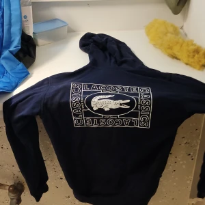 Mörkblå hoodie från Lacoste med tryck - nypris cirka 1300 ja vill bara bli av med den. Snygg mörkblå hoodie från Lacoste med stort vitt tryck på ryggen och mindre tryck på bröstet. Klassisk huva med snörning och känguruficka framtill. Perfekt för dig som gillar streetwear och vill ha en stilren look med tydlig logga.