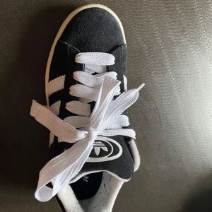 Svarta Adidas campus sneakers - Klassiska svarta Adidas Gazelle sneakers i mocka med vita detaljer och de ikoniska tre ränderna på sidan. Skorna har vit sula, rund tå och breda vita skosnören. Perfekta för en sportig och stilren look.