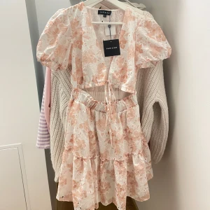Blommig kortklänning från Nelly.com!  - Supersöt kortklänning från nelly.com med broderade blommor i ljusa persikotoner på vit botten. Klänningen har puffärmar, v-ringning, knytning framtill och volangkjol. Perfekt för sommarens alla tillfällen, aldrig använd bara testad. köparen står alltid för frakt!🌸