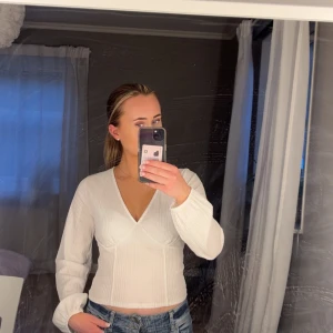 Vit omlottopp med v-ringning - Säljer en vit omlottopp från Gina Tricot med v-ringning och lång ärm. Toppen har en lätt ribbad struktur och sitter figurnära över kroppen. Perfekt att styla med jeans eller kjol för en clean look.