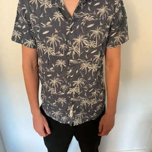 Blå kortärmad skjorta från 157 - Snygg blå skjorta från 157 med korta ärmar och vitt palmträdsmönster. Använd ett fåtal gånger så är myckte bra skick!