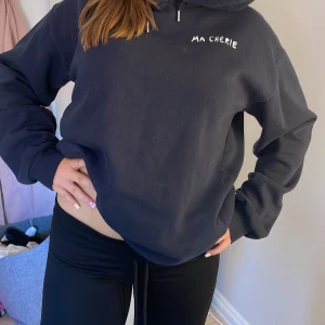Svart hoodie med tryck Ma Chérie - Mörkblå hoodie med vit text 'MA CHÉRIE' tryckt fram och bak, samt två hjärtan på ryggen. Tröjan har huva och en loose passform, perfekt för en chill och avslappnad stil. Materialet känns mjukt och skönt, troligen bomull eller polyester.