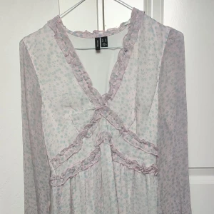 Blommig volangklänning från Vero Moda - Supersöt kortklänning från Vero Moda i ljusrosa med små blå blommor och volangdetaljer. Klänningen har långärmade, transparenta ärmar och v-ringning med volangkant. Perfekt för vår och sommar💞