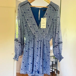 Blå mönstrad kortklänning från Zara - Supersnygg blå kortklänning från Zara med vitt grafiskt mönster. Klänningen har v-ringning, långa transparenta ärmar med resår vid handlederna och markerad midja. Lätt och luftig, perfekt för varmare dagar. Stängs med knapp i nacken.