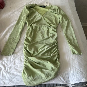 Glittrig grön kortklänning DK Faye - Snygg ljusgrön glittrig kortklänning från DK Faye med omlott-v-ringning, långa ärmar och rynkade detaljer längs sidan. Klänningen har en figurnära passform och är tillverkad i ett glittrigt syntetmaterial som verkligen sticker ut.