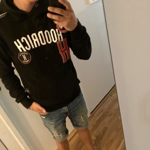 Svart HooDricH hoodie med tryck - Svart hoodie från HooDricH med vit och röd text framtill, patchdetaljer och stor ficka på magen. Tröjan har huva och långa ärmar, perfekt för en chill och sportig look. Snygg streetstyle-vibe och skön passform.
