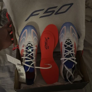 Adidas F50 Elite FG fotbollsskor - Adidas F50 Elite FG fotbollsskor i vitt med blå och röda detaljer. Skorna har snörning, låg profil och en lätt syntetisk ovandel för maxad speed på planen. Snygg design med F50-logga och genomskinlig yttersula med dobbar för gräs. Helt oanvända och nya. Dem passade helt enkelt inte på mig och säljs därför