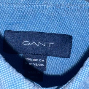 Blå skjorta från GANT, barnstorlek - Snygg blå skjorta från GANT i klassisk modell med button-down krage och vita knappar. Skjortan har lång ärm och broderad GANT-logga på bröstet. Perfekt för dig som vill ha en stilren och tidlös look. Mer ljusblå än mellanblå Stl 134-140, 9-10 år
