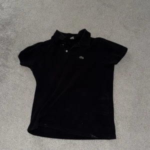 Svart pikétröja från Lacoste - Klassisk svart pikétröja från Lacoste med den ikoniska krokodilloggan på bröstet. Tröjan har korta ärmar, krage och knäppning framtill. Perfekt för en clean och sportig look.