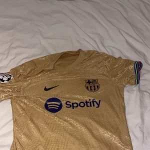 Barcelona guld fotbollströja Pedri #8 - Snygg Barcelona fotbollströja i guld med blå detaljer, Nike-logga och Spotify som sponsor. Tröjan har korta ärmar med regnbågsfärgade muddar och Champions League-märke på ärmen. Pedri och nummer 8 tryckt på ryggen. Materialet är lätt och andas, perfekt för match eller träning.