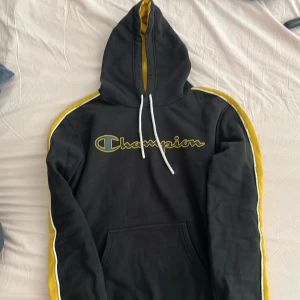 Svart och gul Champion hoodie - Svart hoodie från Champion med gula och vita ränder längs ärmarna och insida av huvan. Stor broderad Champion-logga på bröstet och klassisk känguruficka framtill. Mjuk bomullskänsla och dragsko i huvan. Perfekt för en sportig och chill stil.