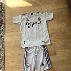 Real madrid tröja - Säljer ett Real Madrid matchställ med tröja och shorts från Adidas i storlek 152. Tröjan är vit med lila detaljer, klubbmärke, sponsortryck och Benzema 9 på ryggen. Shortsen är vita med lila ränder och klubbmärke. Tillverkad i lätt och ventilerande material.