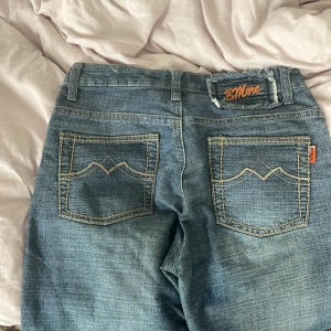 Blå bootcut jeans från BMore - Säljer ett par klassiska blå bootcut jeans från BMore med snygga kontrastsömmar och broderade bakfickor. Jeansen har normal midja, fem fickor och stängs med dragkedja och knapp. Perfekta för dig som gillar en retro vibe och vill ha ett par jeans med lite extra attityd.