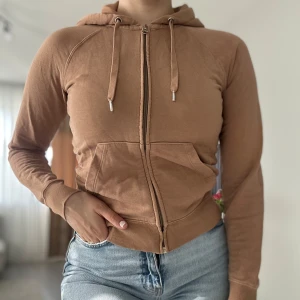 Brun croppad hoodie med dragkedja - Snygg brun hoodie med dragkedja framtill och huva med snörning. Modellen är croppad och har långa ärmar samt ribbade muddar. Perfekt för en chill och avslappnad stil. Praktiska fickor framtill och mjukt bomullsmaterial.