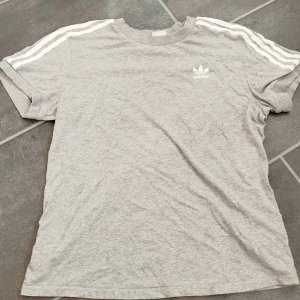 Grå Adidas t-shirt med vita ränder - Snygg grå t-shirt från Adidas med klassiska vita ränder längs axlarna och broderad logga på bröstet. T-shirten har rund hals och korta ärmar, tillverkad i mjuk bomull för en skön känsla. Perfekt för en sportig och avslappnad stil.