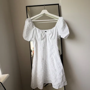Vit blommig kortklänning H&M Divided - Supersöt vit kortklänning med ljusblått blommönster från H&M Divided. Klänningen har puffiga korta ärmar, smock i ryggen och en söt, lätt v-ringad. Använd 1 gång