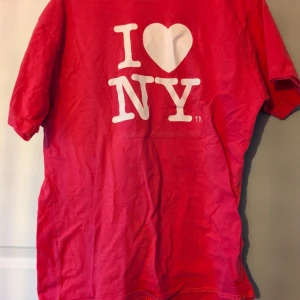 Röd/rosa I Love NY t-shirt - Röd/riaat t-shirt med klassiskt 'I ♥ NY'-tryck i vitt framtill. Skön passform med korta ärmar och rund hals. Perfekt för dig som vill visa lite kärlek till New York. Tillverkad i mjuk bomull som känns bekväm hela dagen.