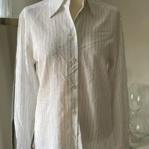 Vit randig skjorta - Snygg vit skjorta med tunna svarta ränder och klassisk krage. Skjortan har långa ärmar och knappar framtill. Perfekt för en stilren look. Lite längre skjorta 