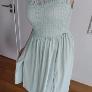 Guess Ljusgrön ärmlös klänning - Säljer en söt ljusgrön klänning med ett diskret geometriskt mönster på överdelen. Klänningen är ärmlös och har en luftig kjol med en liten slits framtill. Perfekt för varma dagar!