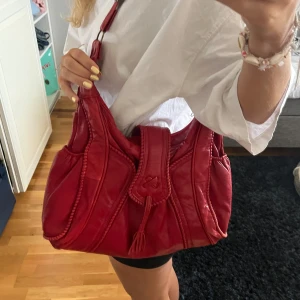 Röd axelväska i skinn - Snygg röd axelväska i skinn med dekorativa sömmar och tofsdetalj. Väskan har flera fack med dragkedja för praktisk förvaring. Perfekt för att ge en färgklick till din outfit.❤️❤️🪩