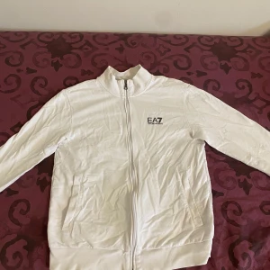 Vit jacka från Emporio Armani - Säljer en stilren vit jacka från Emporio Armani med dragkedja och hög krage. Jackan har långa ärmar och en broderad logotyp på bröstet. Perfekt för en sportig look! Behöver bara strykas, Storlek S