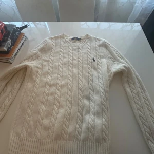 Vit stickad tröja från Ralph Lauren - Säljer en stilren vit stickad tröja från Ralph Lauren med kabelstickat mönster och den klassiska loggan på bröstet. Tröjan har långa ärmar och ribbade muddar vid ärmslut och nederkant. Perfekt för kyliga dagar!