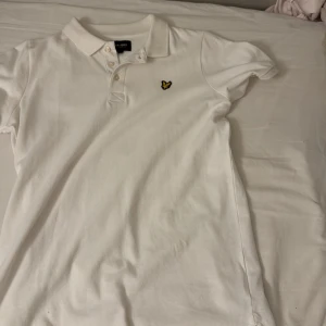 Vit kortärmad pikétröja från Lyle & Scott - Säljer en klassisk vit pikétröja från Lyle & Scott med korta ärmar och en broderad logga på bröstet. Tröjan har en krage med knappar och är perfekt för en stilren look.