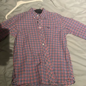 Rutig skjorta från Fred Perry - Snygg rutig skjorta från Fred Perry i rött, blått och vitt. Skjortan har långa ärmar och knappar framtill. Perfekt för en stilren look.