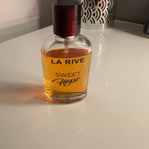 La Rive Sweet Hope Eau de Parfum - Säljer en elegant flaska av La Rive Sweet Hope Eau de Parfum. Flaskan är genomskinlig med en stilren design och har en mörk kork. Doften är varm och inbjudande, perfekt för den som vill ha en söt och hoppfull känsla.