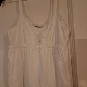 Vit spetsprydd linnetopp från Nelly - Säljer en söt vit linnetopp från Nelly med spetsdetaljer vid halsringningen och axelbanden. Toppen har en v-ringad design och en liten rosett framtill. Perfekt för en somrig look!