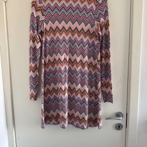 Mönstrad klänning från Gina Tricot - Säljer en färgglad klänning från Gina Tricot med ett zigzag-mönster i rosa, lila, orange och svart. Klänningen har långa ärmar och en bekväm passform. Perfekt för att liva upp garderoben! (Katt o kanin finns i hemmet) 