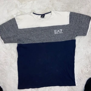 T-shirt från EA7 Emporio Armani passar även L  - Snygg t-shirt från EA7 Emporio Armani i en stilren design med blockfärger. Den har en kombination av vit, grå och mörkblå färg med korta ärmar. Perfekt för en avslappnad stil.